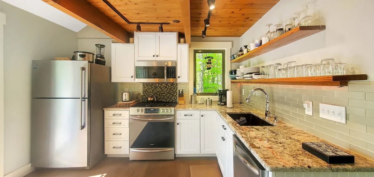 A modern kitchen with a stainless steel refrigerator, granite countertop, and open shelving showcasing neatly arranged glassware. A window allows natural light to spill in, highlighting a space ideal for cooking, hosting, and sharing meals with friends or family.