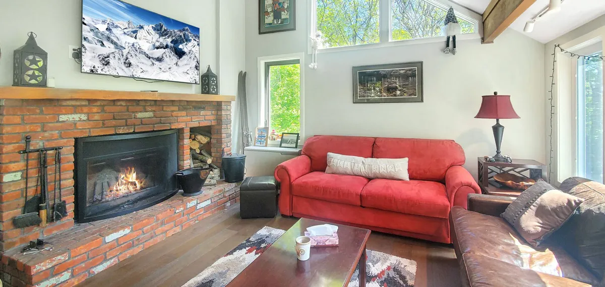A cozy living area dominated by a glowing fireplace, a red couch, and a television. The room exudes warmth and a welcoming atmosphere, ideal for group gatherings, movie nights, or curling up after a day in the snow.