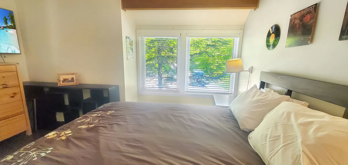 A bedroom with a plush bed adorned in decorative pillows, ample natural light streaming through large windows, a television mounted on the wall, a wooden dresser, and a modern shelving unit. The space feels tranquil and ideal for unwinding.