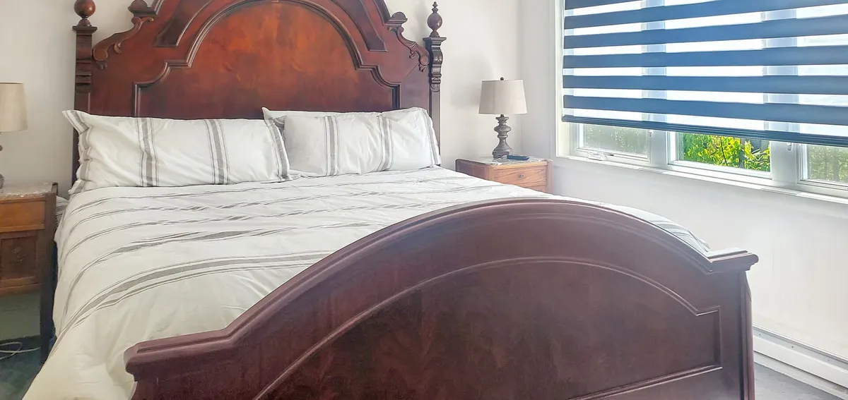 A serene bedroom with an ornate wooden bed frame, striped window blinds allowing gentle natural light, and nightstands with lamps. The space exudes tranquility, perfect for a romantic morning or cozy evening in.