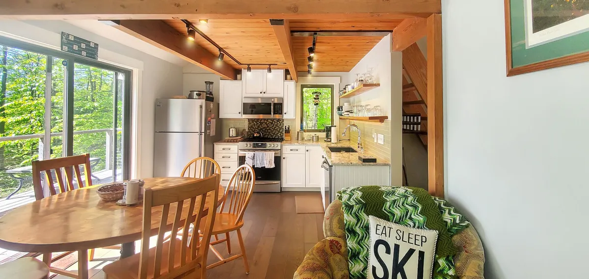 The image showcases a sunlit, open-concept dining and kitchen area with a spacious wooden table set for family gatherings, a modern kitchen perfect for group meals, and a plush armchair draped with a cozy blanket, inviting guests to unwind after a day on the slopes.
