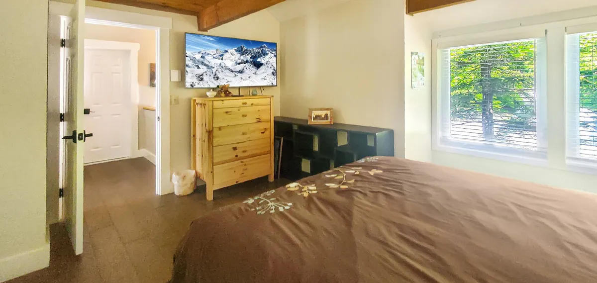 A bright bedroom featuring a bed with floral embroidery, a television displaying a mountain landscape, a wooden dresser, and large windows that look out onto lush greenery. The room feels comfortable and airy, inviting relaxation and leisure.