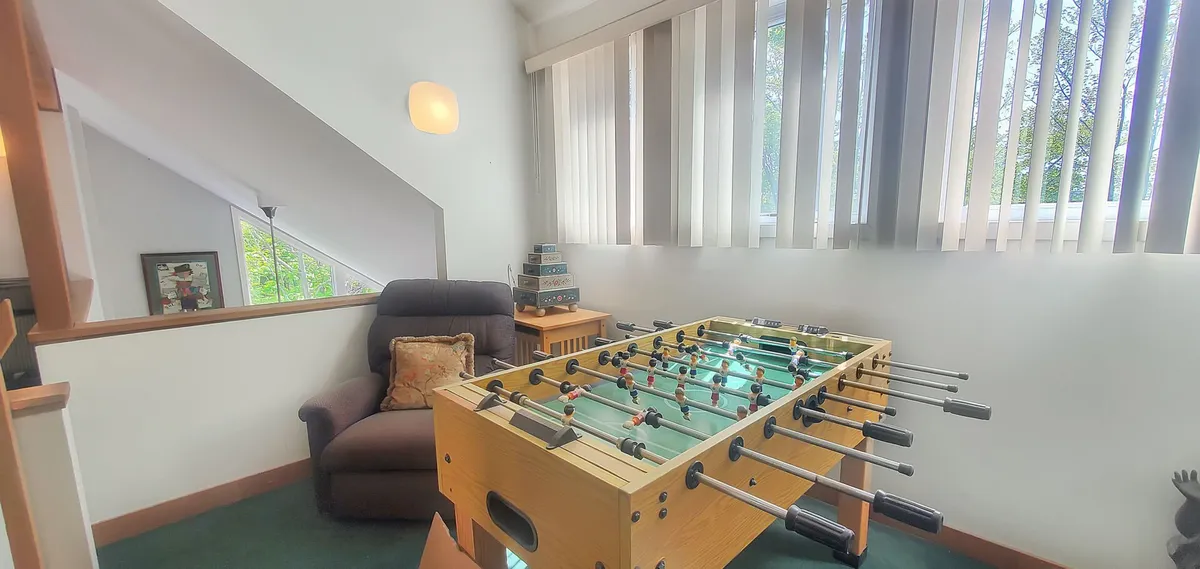 This image features a foosball table as the central element, complemented by a comfortable chair and large windows with blinds that let in natural light. The overall vibe is casual and inviting, perfect for family bonding and friendly competition.