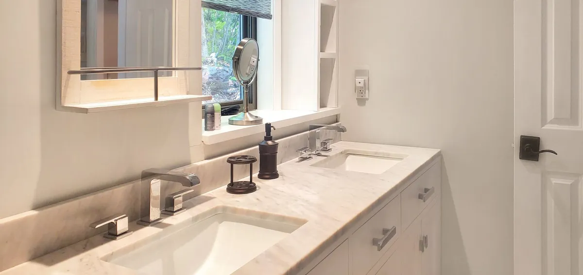 A sparkling clean bathroom with a double sink, marble countertop, a large mirror, and a window offering a glimpse of greenery. The space feels fresh, functional, and soothing—ideal for morning routines or evening wind-downs.