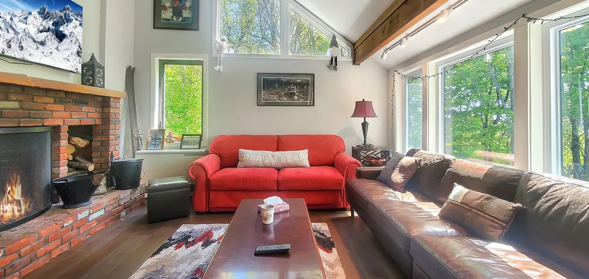 A welcoming living room featuring a plush red sofa positioned near a substantial brick wood-burning fireplace. Large windows frame views of nature and fill the space with natural light, while a coffee table sits at the center—perfect for board games, hot drinks, and social gatherings.